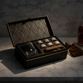 An open black gift box with gold geometric leaf patterns on the inside lid, containing a set of small bottles and a dark glass container, placed on a marble surface with a brown velvet pouch beside it.