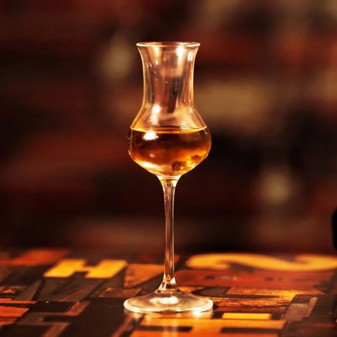 A clear glass tulip-shaped decanter or goblet filled with amber liquid, standing on a dark wooden surface in a warm, dimly lit setting.