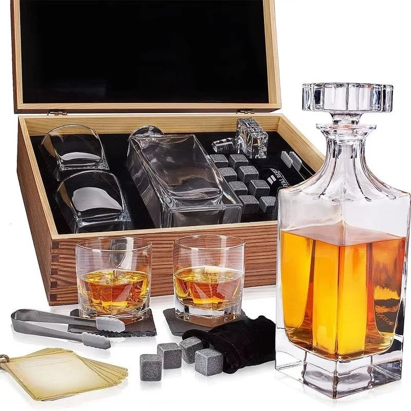 A whiskey decanter gift set displayed with its wooden presentation box open, including a clear glass decanter filled with amber liquor, two small whiskey glasses, a glass crumb catcher, gray ice cubes, metal tongs, a black cloth, and a small card set, all arranged on a white background.
