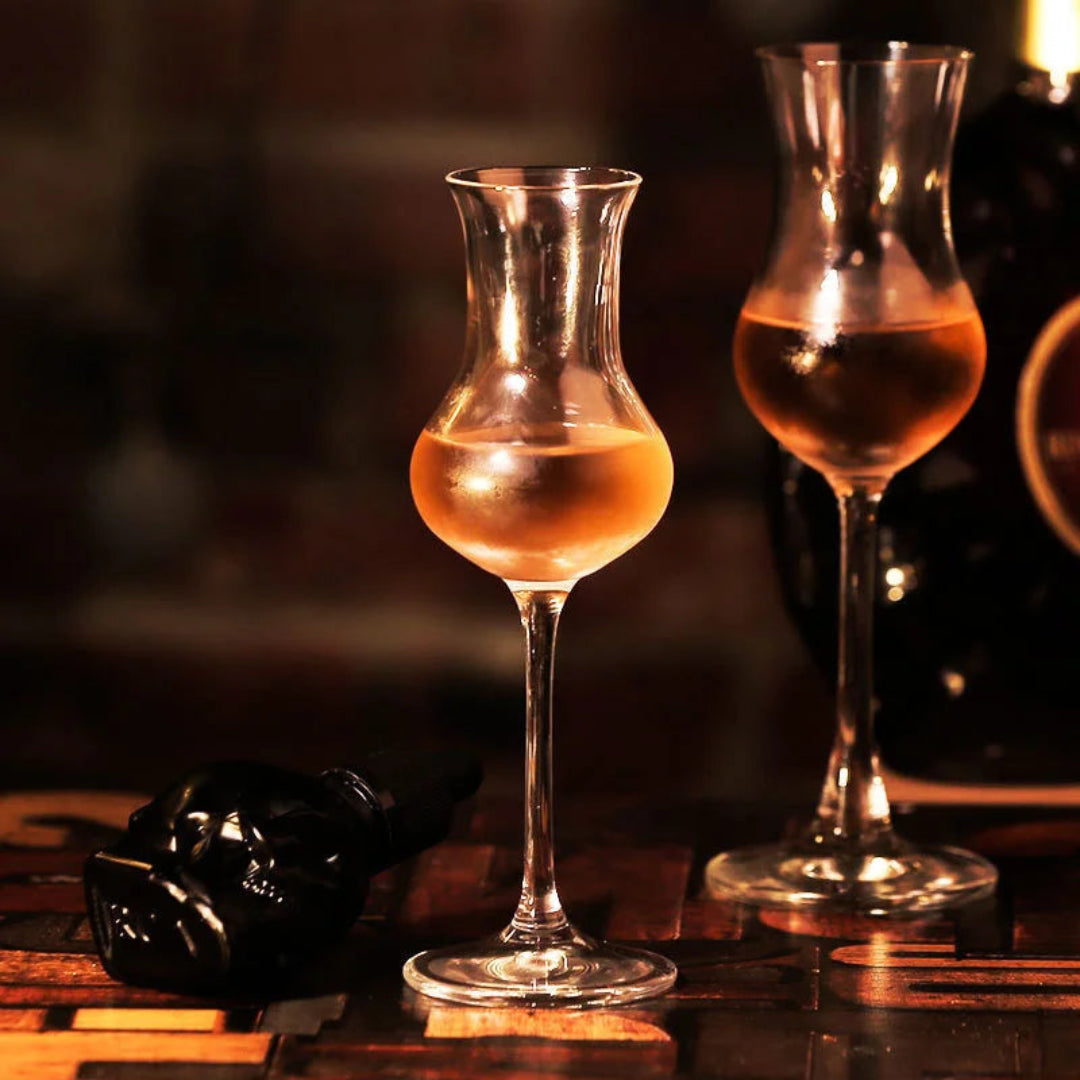 Two clear glass tulip-shaped decanters or goblets filled with amber liquid, set on a dark wooden table in warm, dim lighting.