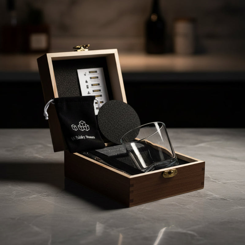 A wooden presentation box opened on a stone countertop, containing a clear glass, black textured accessories, a black drawstring pouch with white 'Whisky Stones' branding, and a white card insert. The background is a softly blurred dark kitchen or bar setting.