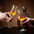 Two hands holding tulip-shaped crystal wine glasses filled with amber liquor, clinking together in front of a dark brown background with a bottle and another glass partially visible.