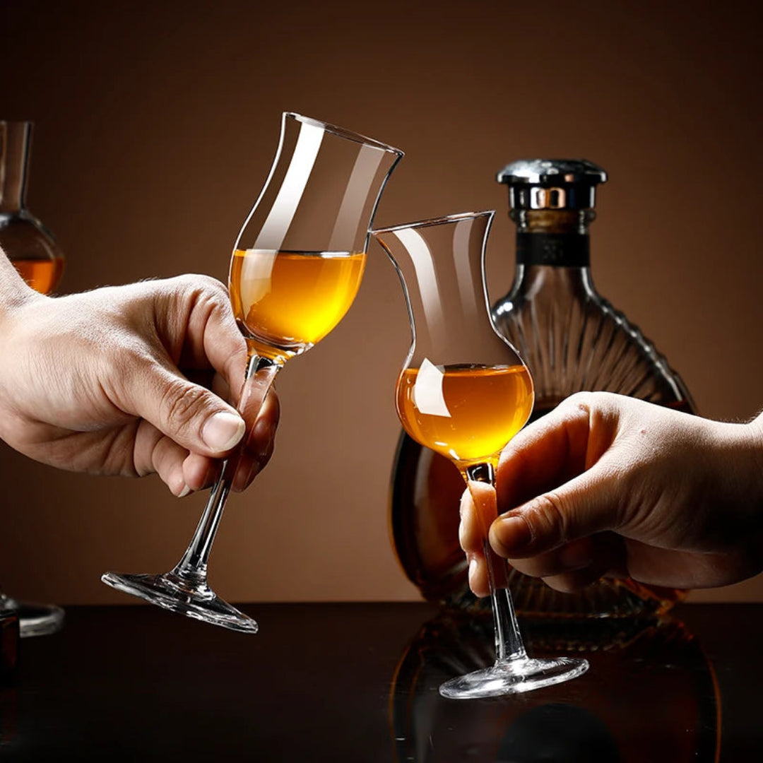 Two hands holding tulip-shaped crystal wine glasses filled with amber liquor, clinking together in front of a dark brown background with a bottle and another glass partially visible.