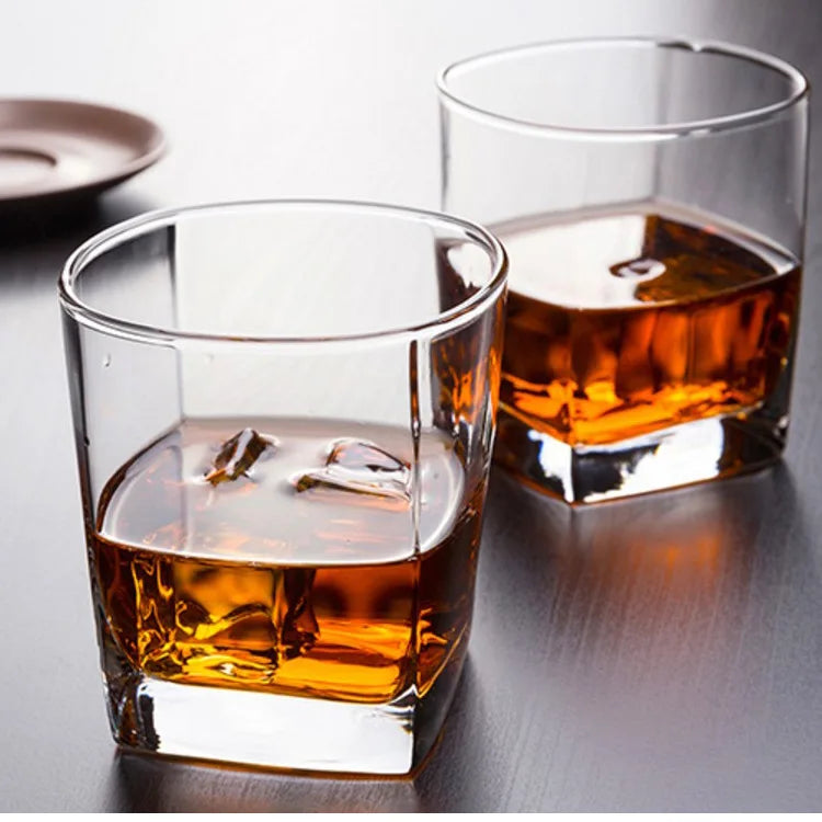 Two clear square rocks glasses filled with amber liquor and ice, photographed on a dark tabletop with a softly blurred background.